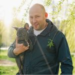 Mand holder fransk bulldoghvalp i solbeskinnet park, smilende